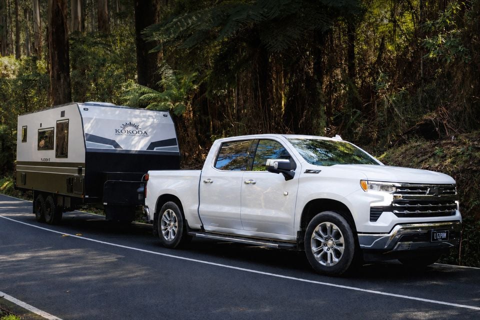 A white Chevrolet Silverado LTZ crew-cab ute towing a Kokoda caravan along a forested road