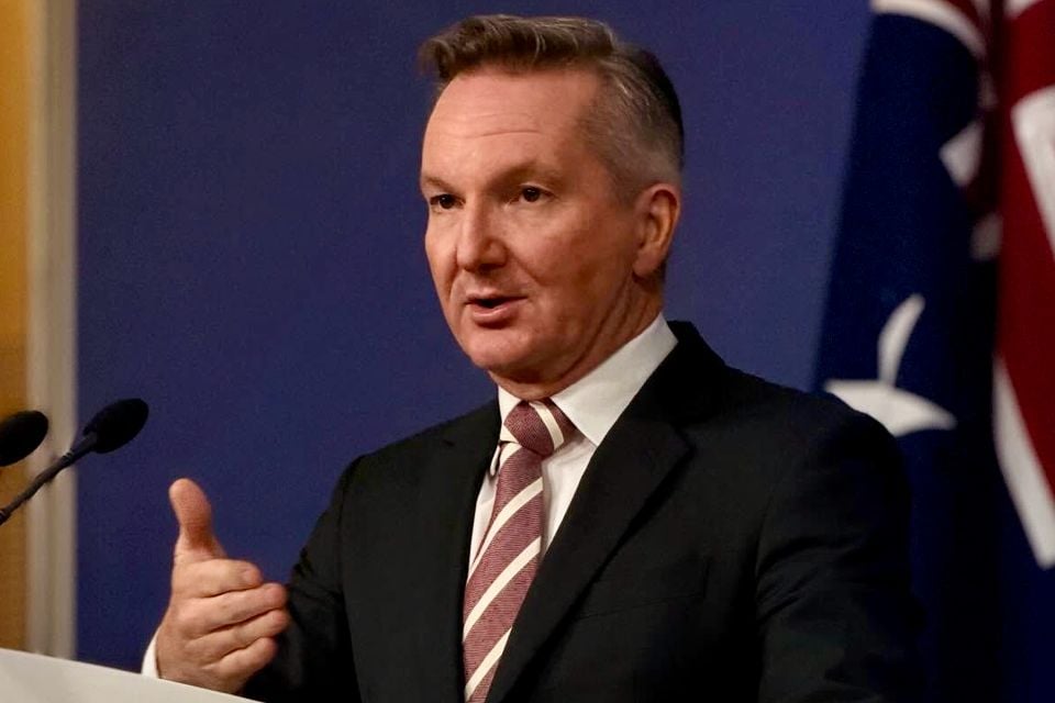 Chris Bowen de terno escuro e gravata listrada falando em um púlpito com microfones, uma bandeira australiana atrás dele