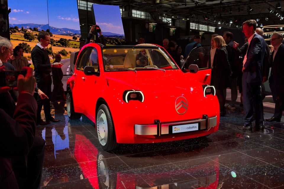 Um carro-conceito Citroen 2CV vermelho com a porta aberta, cercado por curiosos e fotógrafos em um estande do salão do automóvel