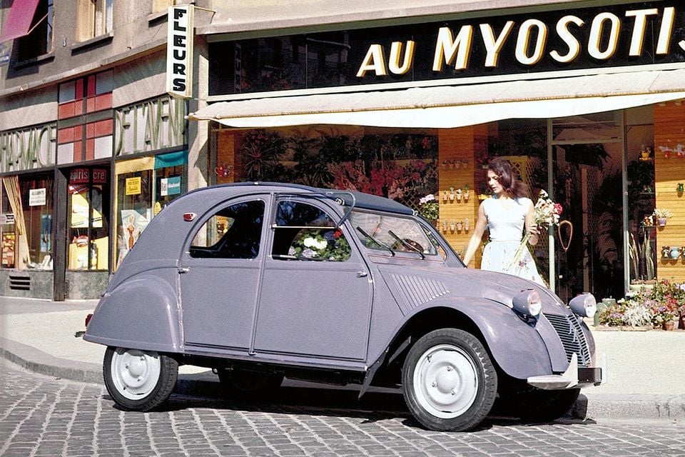 A grey vintage Citroen 2CV parked on a cobblestone street outside a florist, with a woman holding flowers nearby