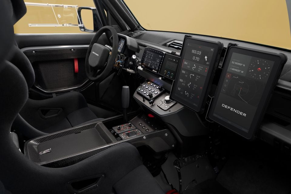 Interior of a Land Rover Defender rally car showing carbon fibre trim, racing seats, a rally-spec centre console with toggle switches, and dual large navigation screens mounted on the dashboard