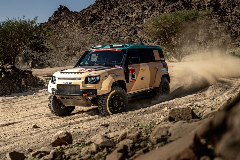 A Land Rover Defender rally car (number 502) in desert-sand livery racing on a dusty dirt track through rocky terrain
