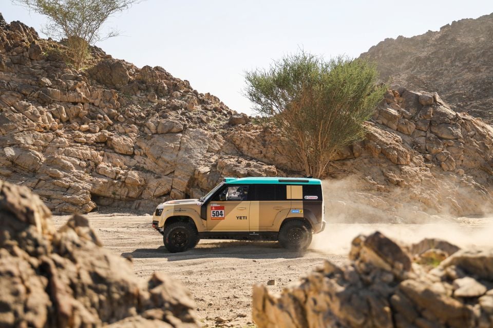 A Land Rover Defender rally car numbered 504 with 'Yeti' livery driving through a rocky desert landscape, kicking up dust