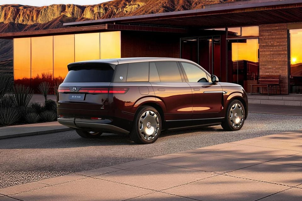A dark red Denza N9 SUV parked outside a modern building at sunset, seen from the rear and side with mountains behind