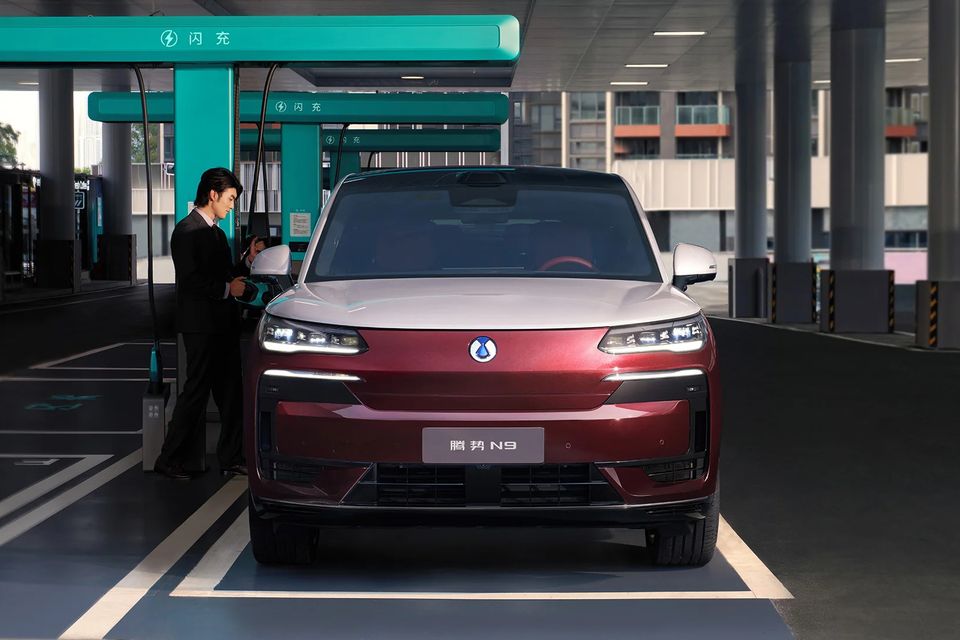 A red and silver Denza N9 SUV seen from the front at a charging station, with a man plugging in the charging cable