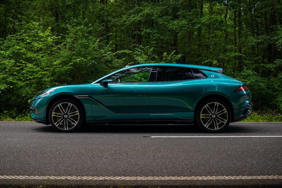A teal Denza Z9 GT wagon seen from the side, parked on a road in front of lush green forest