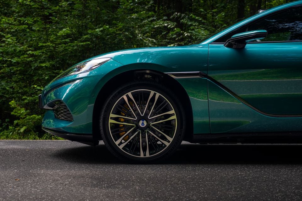 Close-up of the front wheel and side profile of a teal Denza Z9 GT, showing a large multi-spoke alloy wheel with yellow brake callipers, parked on a road with trees in the background.