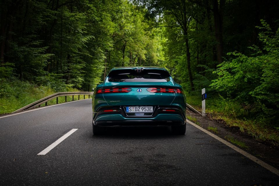 A teal Denza Z9 GT seen from the rear, driving along a tree-lined road through a forest