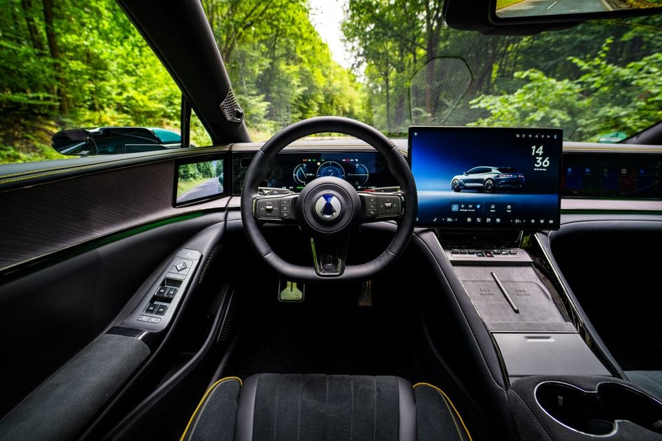 Interior of the Denza Z9 GT showing the dashboard, steering wheel with brand logo, large central touchscreen infotainment display, digital instrument cluster, and alcantara seats with yellow trim stitching