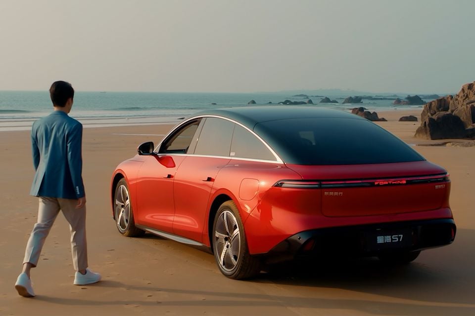 A red Dongfeng Forthing S7 sedan seen from the rear three-quarter angle, parked on a beach with a person walking towards it