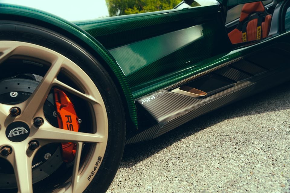 Close-up of the Donkervoort P24 RS showing its green carbon fibre sill, carbon side skirt badged P24 RS, gold alloy wheel with orange brake caliper, and red racing seat visible through the open door