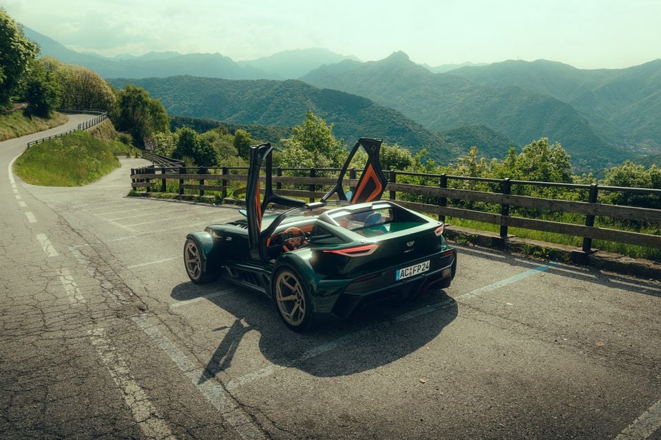 A dark green Donkervoort P24 RS supercar with scissor doors open, seen from the rear three-quarter angle on a mountain road with a scenic valley backdrop.