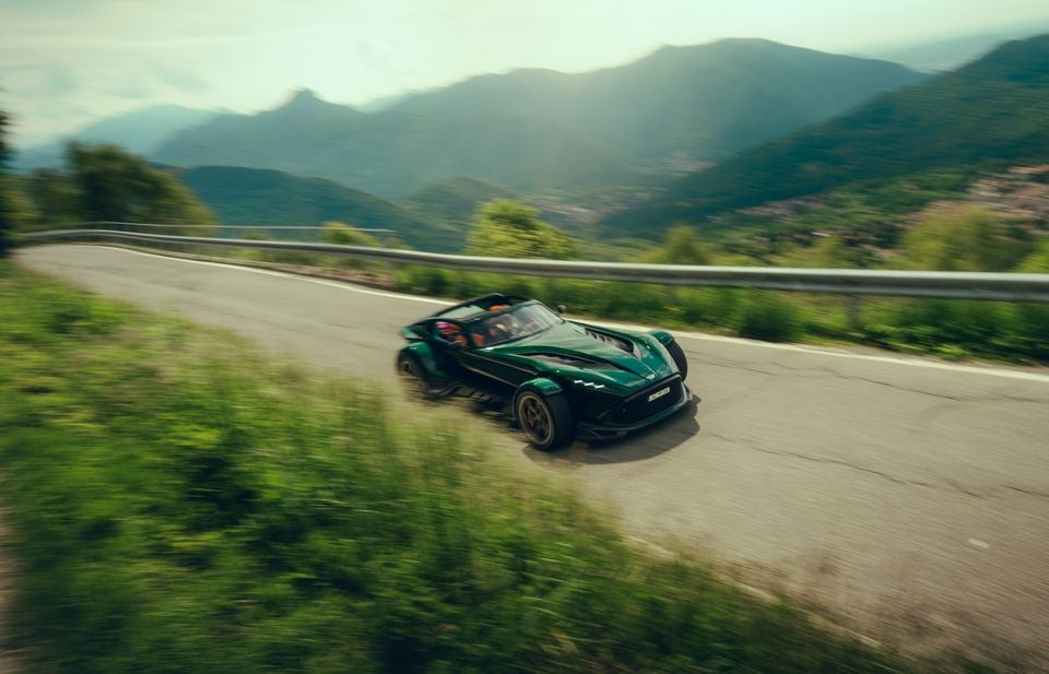 A dark green Donkervoort P24 RS open-top sports car driving along a mountain road, seen from the front-side