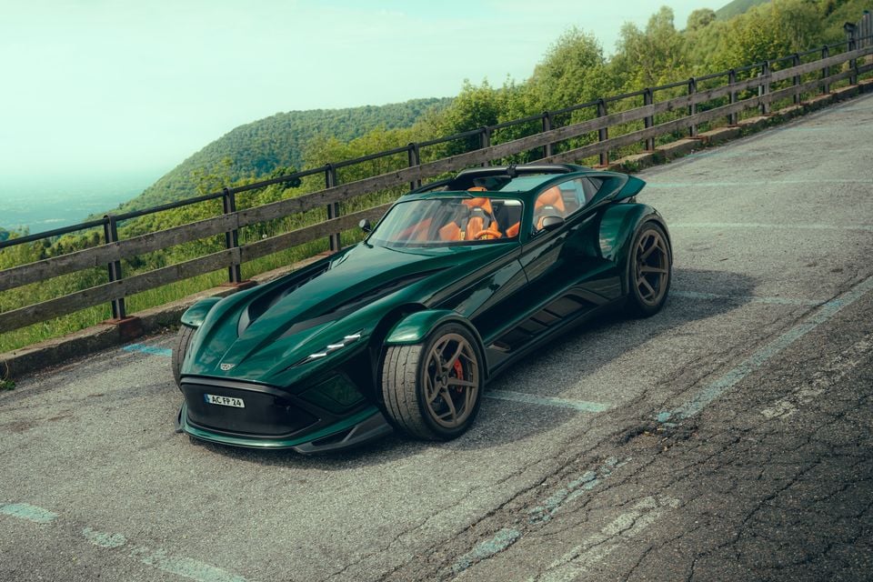 A dark green Donkervoort P24 RS open-top sports car seen from the front-side, parked on a hilltop road with green hills in the background