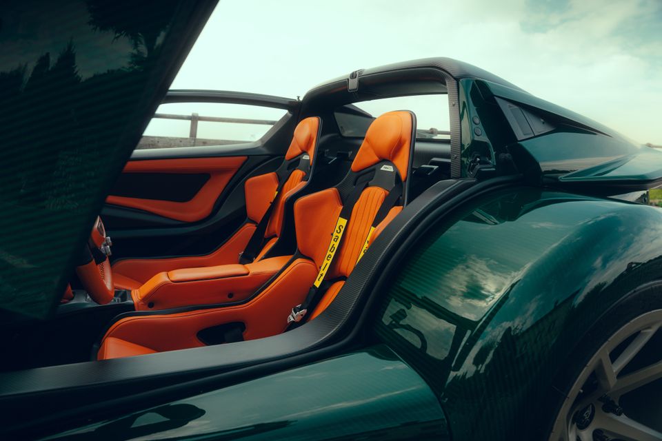 Interior of a Donkervoort P24 RS supercar showing two orange leather bucket seats with racing harnesses and carbon fibre trim, viewed through the open door of the dark green body