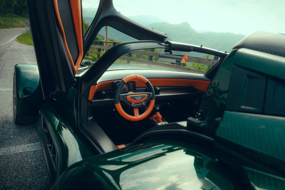 Interior of a Donkervoort P24 RS with the scissor door open, showing an orange leather steering wheel, carbon fibre trim, and bucket seats, with a mountain road visible in the background