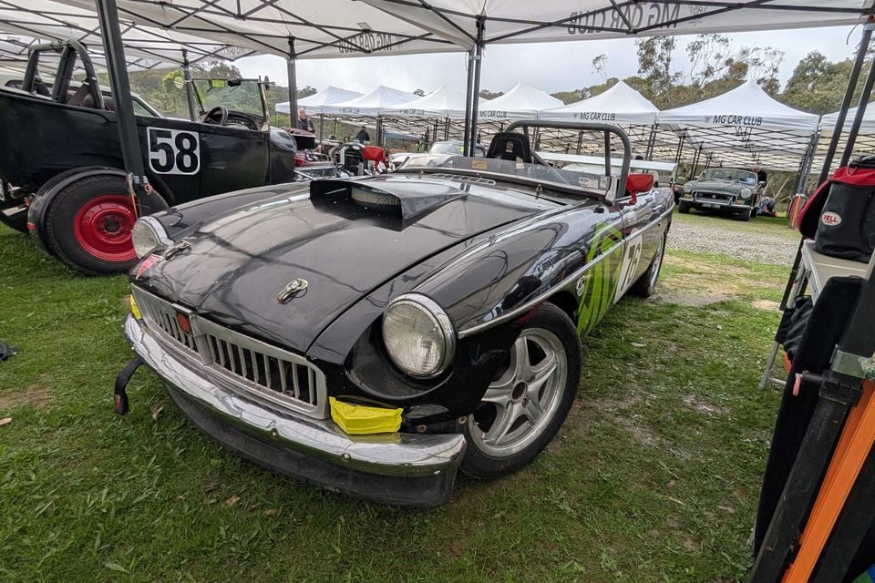 A black MGB roadster race car with a bonnet scoop and roll bar parked on grass under MG Car Club marquees at a motorsport event