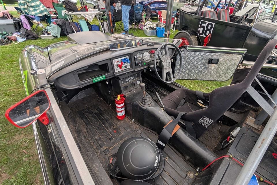 Stripped-out cabin of an MGB V8 race car with a bucket seat, harness, steering wheel, fire extinguisher and helmet on the floor