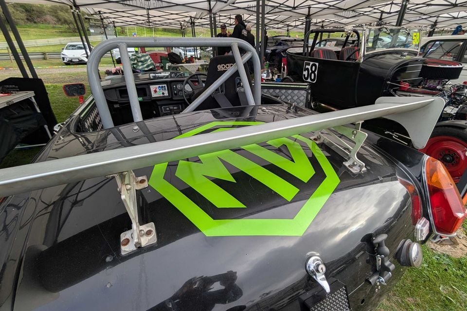 Black MGB V8 race car seen from behind, with a large green MG logo on the boot, a rear wing and a roll cage, parked in a racetrack paddock