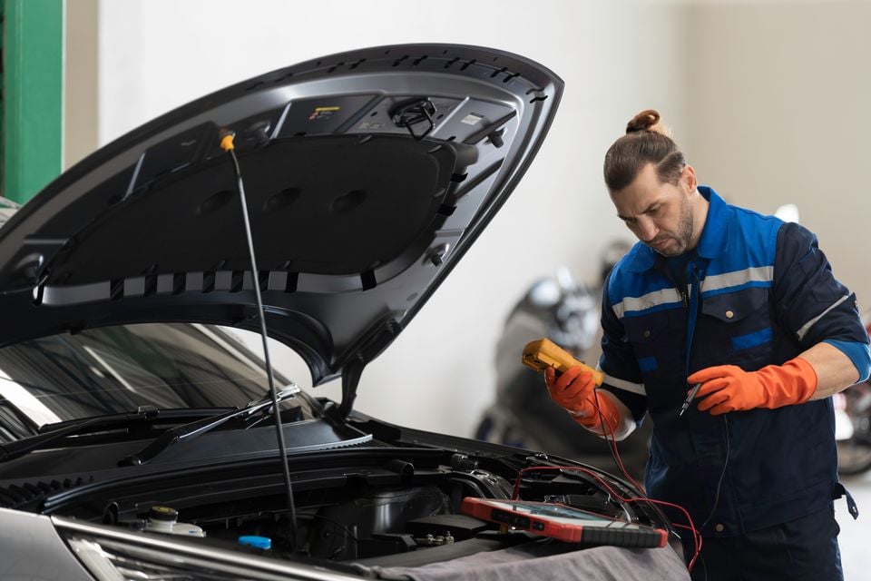 A mechanic in blue overalls and orange gloves uses a multimeter to test a car battery with the bonnet open in a workshop.