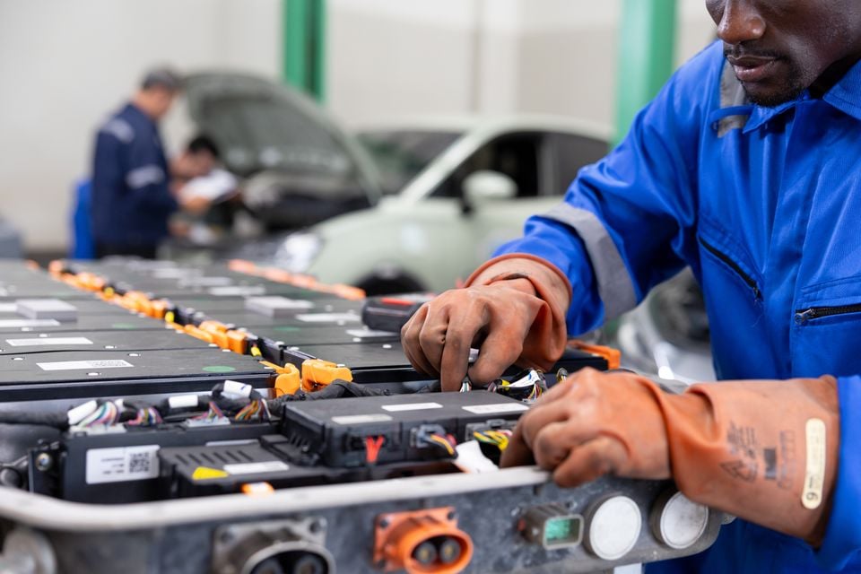 A mechanic wearing rubber gloves works on an electric vehicle battery pack in a workshop, with other technicians and a car visible in the background.