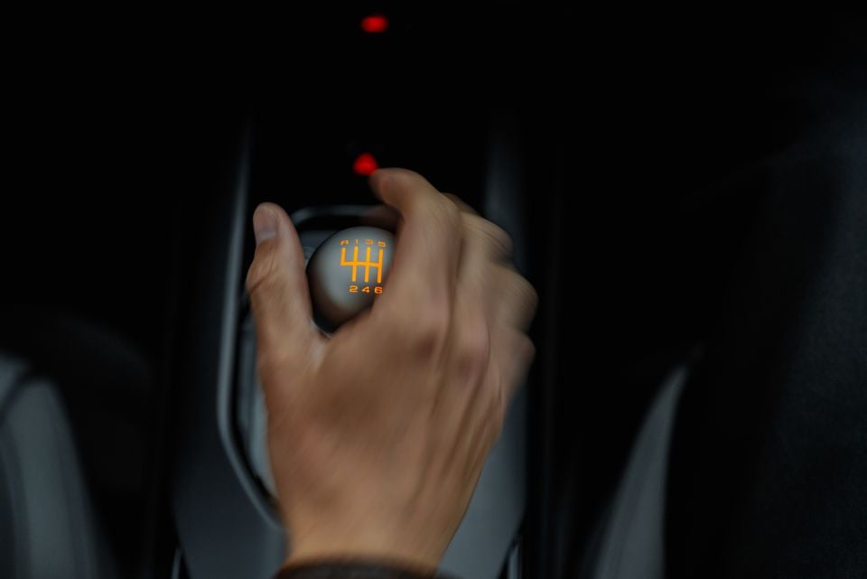A hand gripping a 6-speed manual gear shift knob with an orange shift pattern diagram inside a car interior