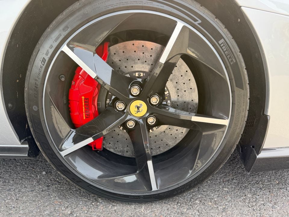 Close-up of a Ferrari wheel showing a red Brembo carbon ceramic brake caliper, drilled disc rotor, five-spoke black alloy wheel with Ferrari prancing horse centre cap, and Pirelli tyre