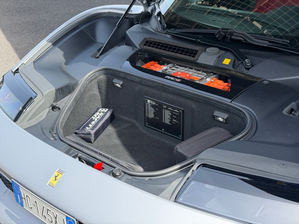 The front trunk (frunk) of a Ferrari open to reveal a small carpeted storage compartment with a safety kit and information plaque, with high-voltage EV components visible behind