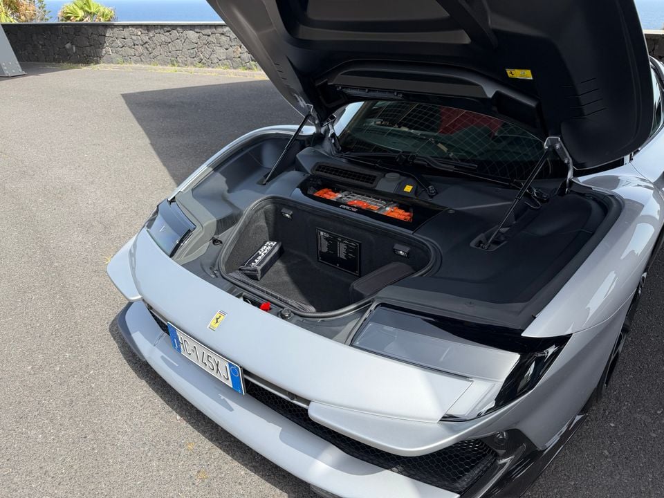 The open front storage compartment (frunk) of a silver Ferrari, showing luggage space and components beneath the raised hood