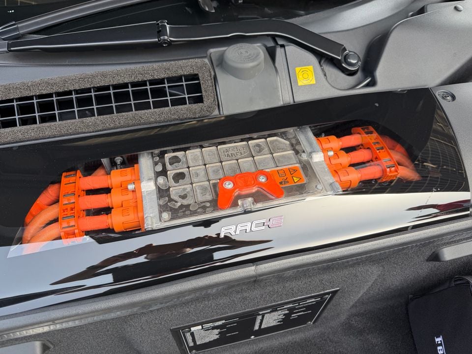 Close-up of the Ferrari hybrid battery unit with orange high-voltage cables and connectors, labelled RAC-E, visible under an open engine cover
