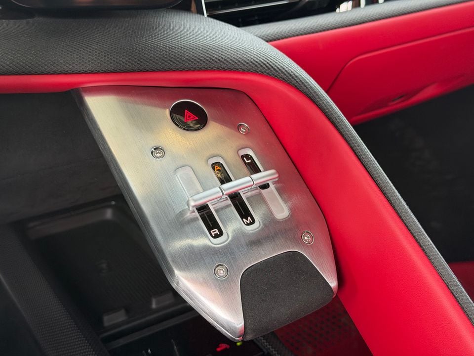 Close-up of the gear selector panel on a Ferrari interior, featuring a brushed aluminium surround with automatic, manual and launch mode switches, set against red leather upholstery