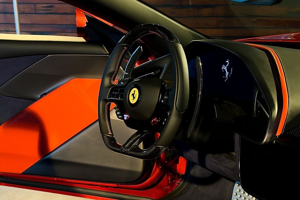 Ferrari interior showing a flat-bottomed carbon fibre steering wheel with the prancing horse logo, orange and black door trim, and a digital display showing the Ferrari badge