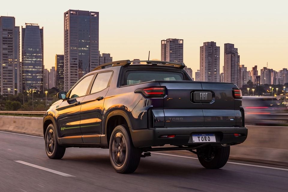 A Fiat Toro ute seen from the rear three-quarter angle driving on a highway with a city skyline in the background at dusk