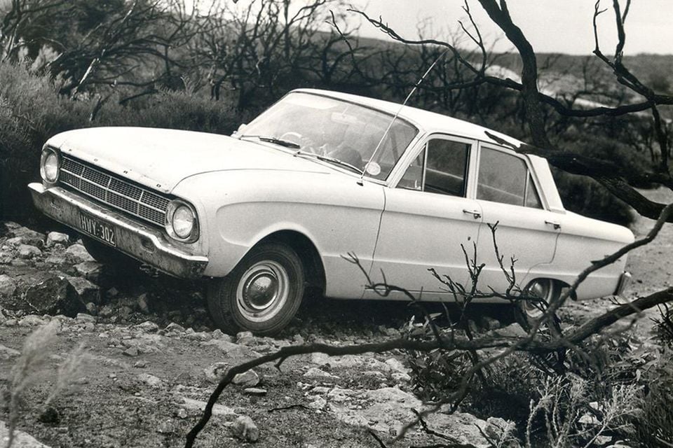 A vintage white Ford Falcon XM sedan parked on rough rocky ground amid dead bushes, photographed in black and white during testing.