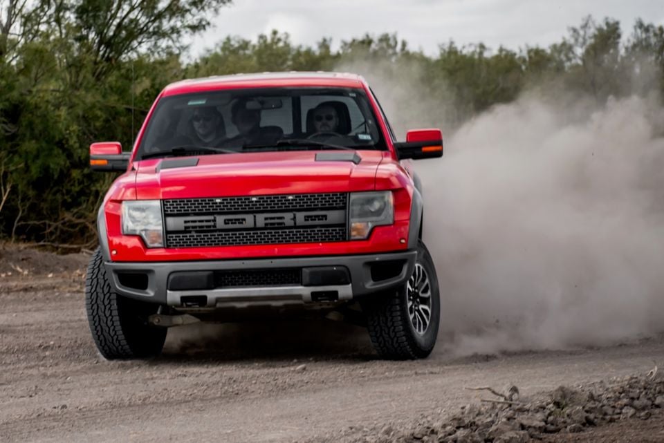 A red Ford F-150 SVT Raptor ute driving fast on a dirt track, kicking up a large dust cloud, seen from the front