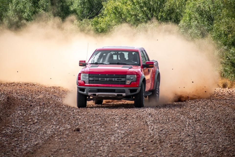 A red Ford F-150 SVT Raptor ute driving aggressively on a dirt track, kicking up a large cloud of dust, seen from the front