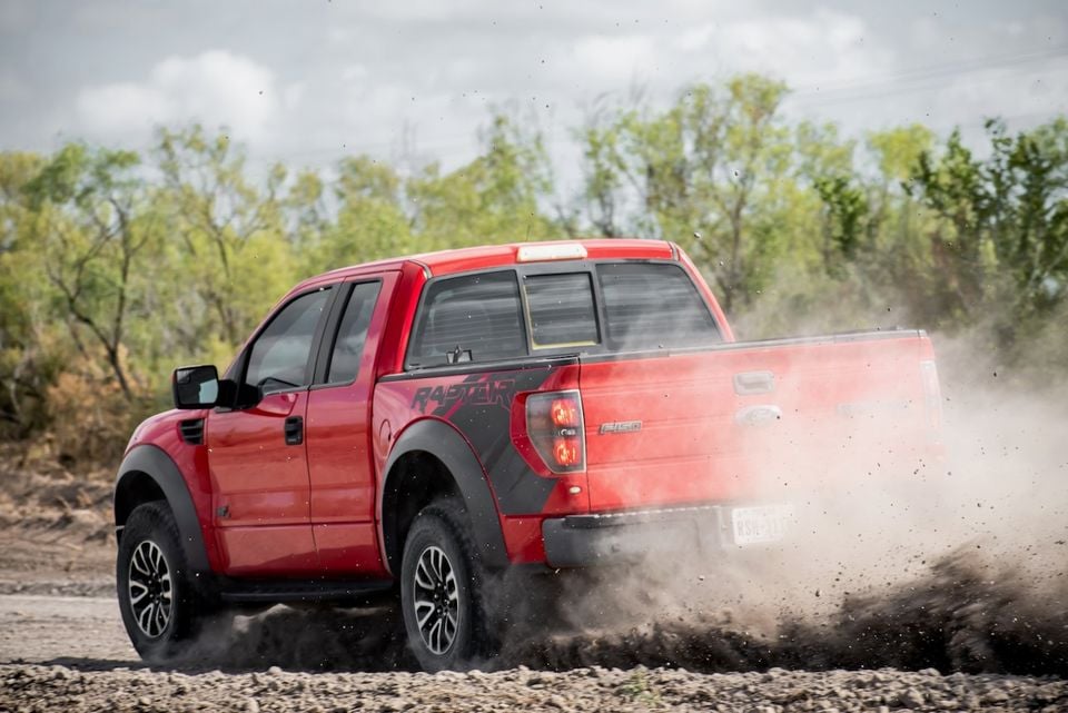 A red Ford F-150 SVT Raptor ute kicking up dirt and gravel on an off-road track, seen from the rear three-quarter angle