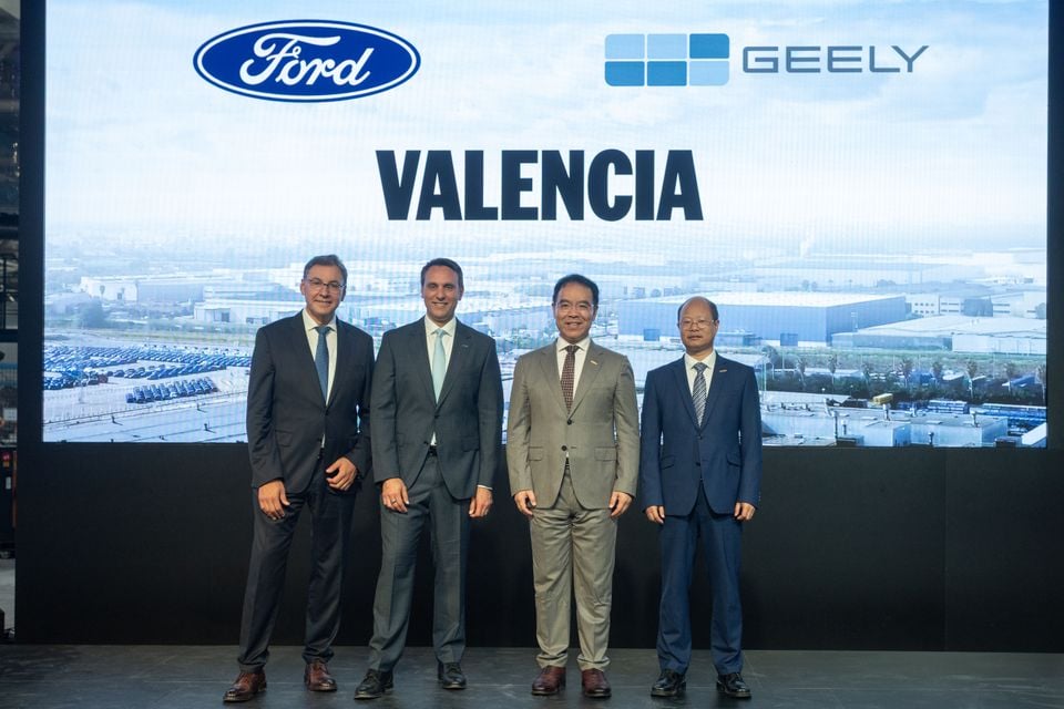 Ford and Geely executives pose together in front of a large screen displaying both company logos and the word 'Valencia' at a joint announcement event.