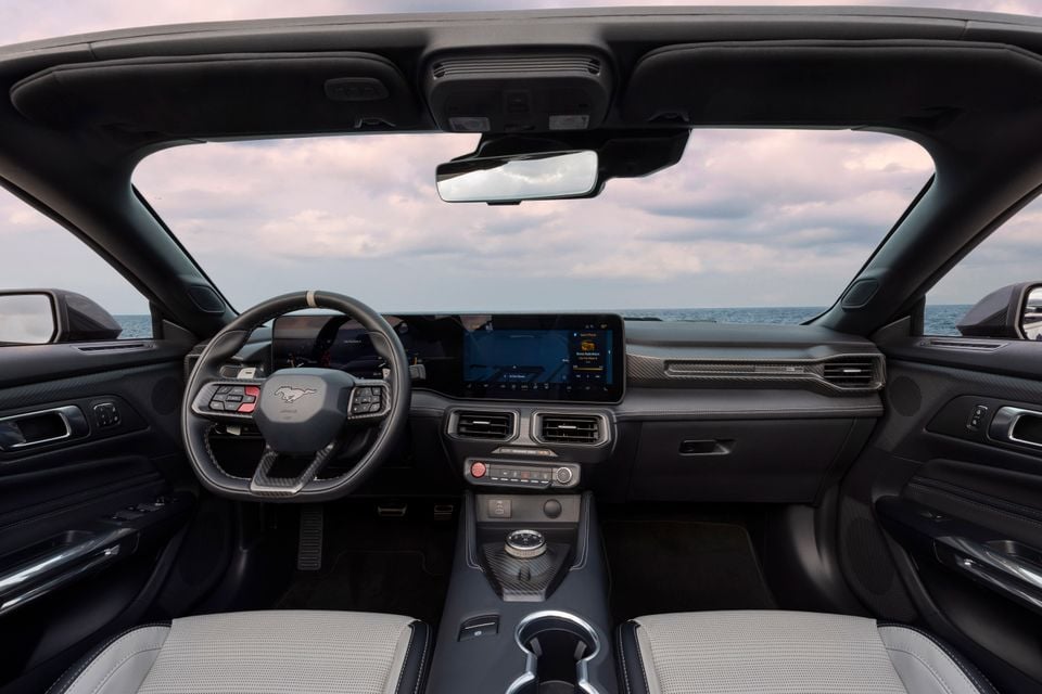 Interior of a Ford Mustang Dark Horse convertible showing the dashboard, Mustang-badged steering wheel, wide touchscreen infotainment display, carbon fibre trim, and light grey front seats with an ocean view through the windscreen