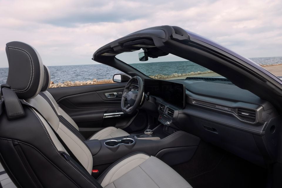 Interior of a Ford Mustang Dark Horse convertible showing two-tone black and grey leather seats, steering wheel, large infotainment touchscreen, centre console and carbon fibre trim, with the top down and ocean visible in the background