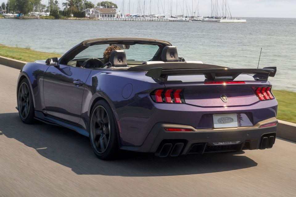 A purple Ford Mustang Dark Horse convertible seen from the rear three-quarter angle, driving along a road beside a marina with the top down.