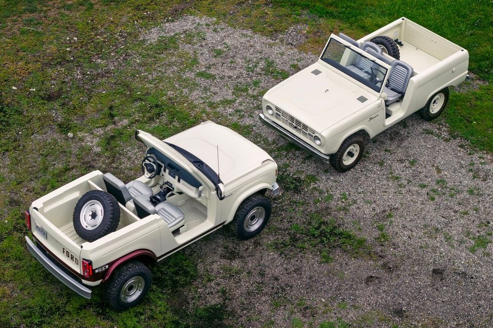 Aerial view of two white Ford Broncos side by side — a modern Ford Bronco Roadster concept with doors and roof removed alongside a classic first-generation Ford Bronco, both finished in white, parked on a gravel and grass surface.