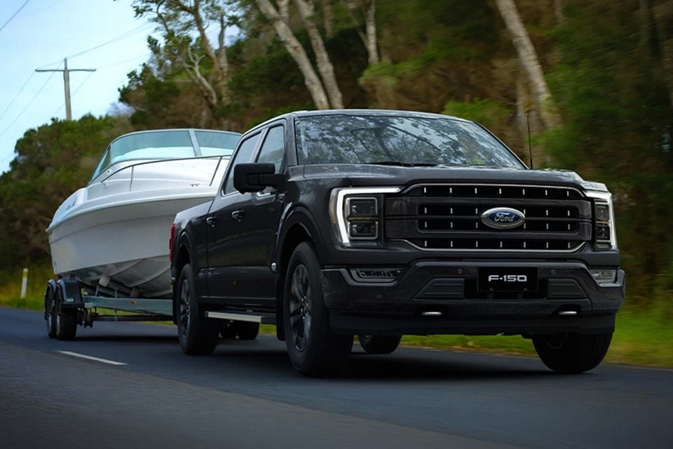 A black Ford F-150 pickup towing a white boat on a trailer along a tree-lined country road, seen from the front