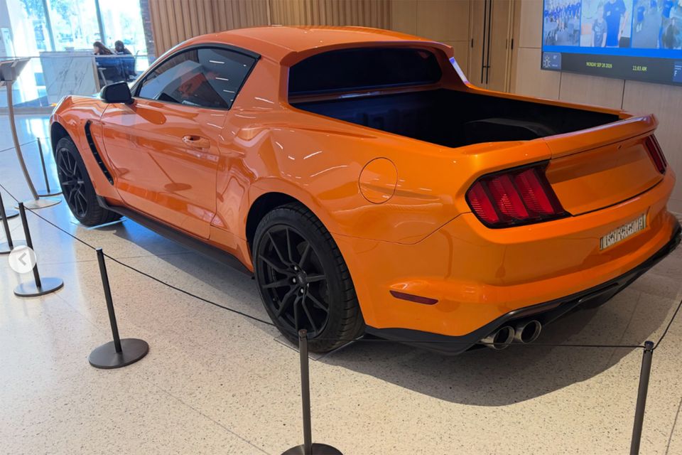 An orange Ford Mustang converted into a ute with a tray at the back, displayed behind rope barriers in a building foyer, seen from the rear