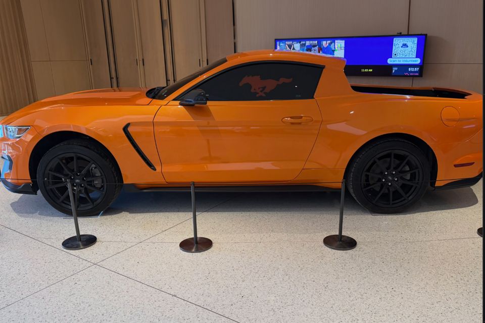 An orange Ford Mustang converted into a ute with an open tray, displayed side-on behind stanchions in an indoor lobby
