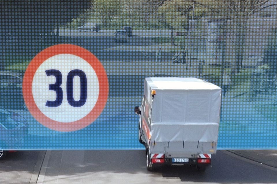 A small delivery van passes in front of a large display showing a 30km/h speed limit sign, illustrating geofencing technology that could automatically limit vehicle speeds.
