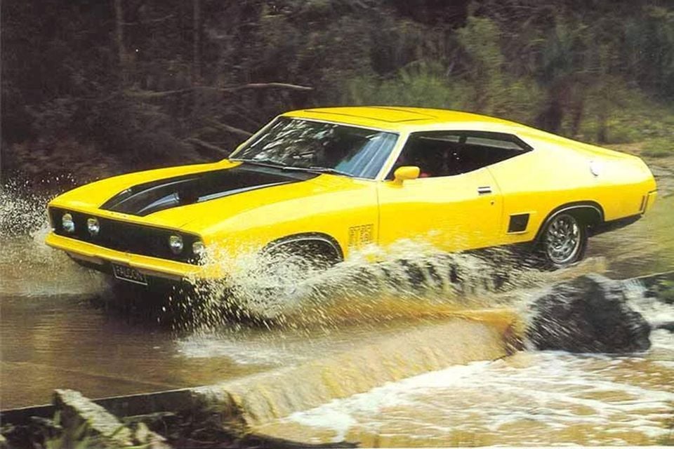 A yellow Ford XB GT Falcon hardtop splashes through a water crossing, viewed from the front quarter