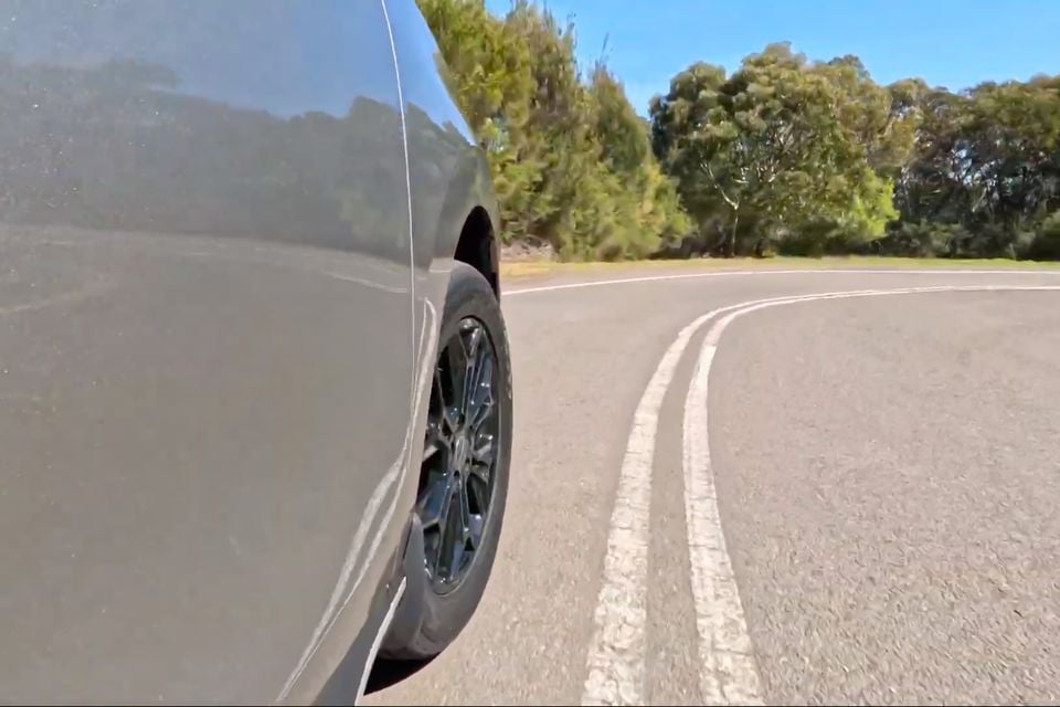 Close-up low-angle shot of a grey car's rear wheel and lower body panel on a country road lined with trees