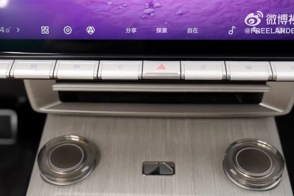 Close-up of a car's centre console showing a large touchscreen infotainment display with physical shortcut buttons below it and two rotary dials on the console panel