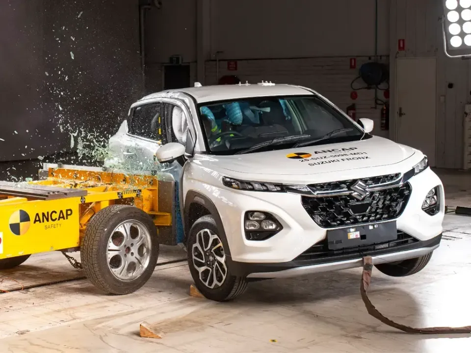 A white Suzuki Fronx SUV undergoing an ANCAP side-impact crash test, with glass shattering from the driver's side window as it strikes the yellow barrier
